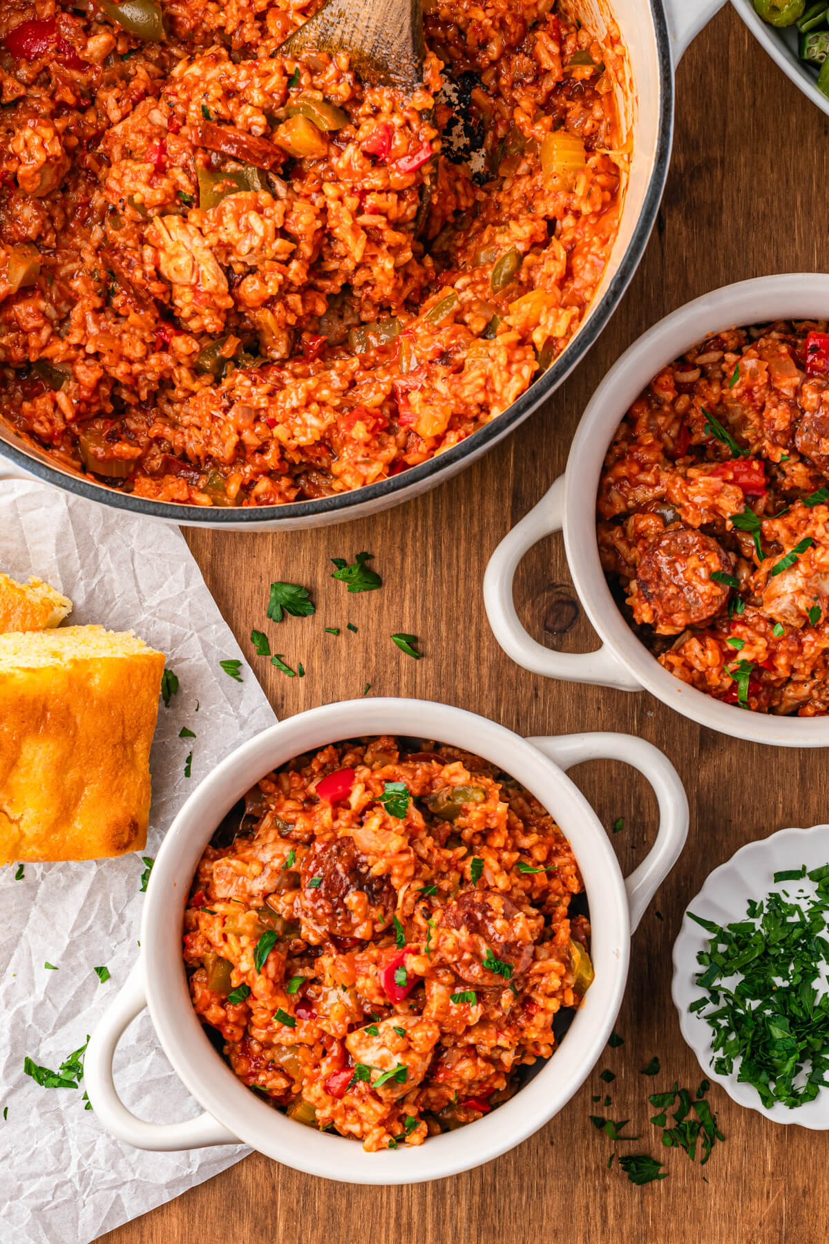 A full Dutch oven beside two bowls of vibrant red sausage chicken jambalaya with red and green bell peppers garnished with fresh parsley.