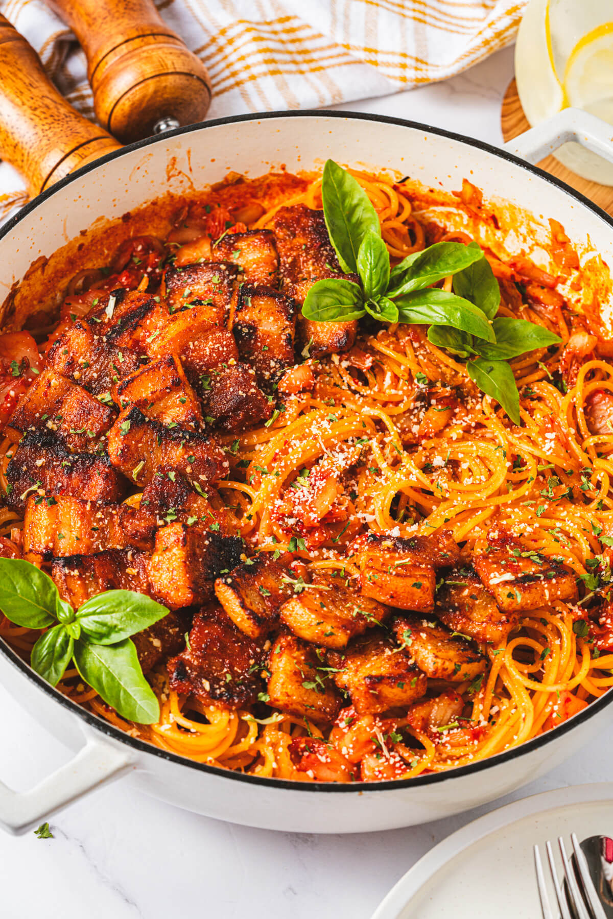A Dutch oven filled with vibrant red Kimchi Pasta dotted with caramelized pork belly and fresh basil leaves.