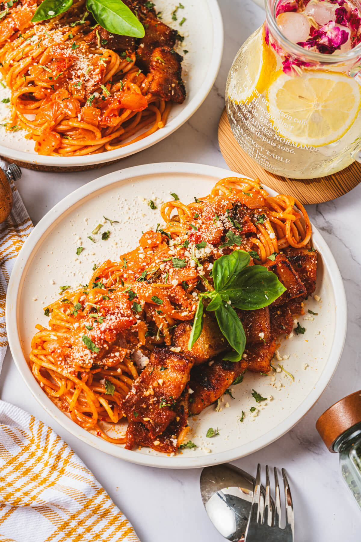 A white dinner plate with a serving of Kimchi Pasta with pork belly expertly rolled into a neat line then topped with Parmesan cheese and fresh basil leaves.