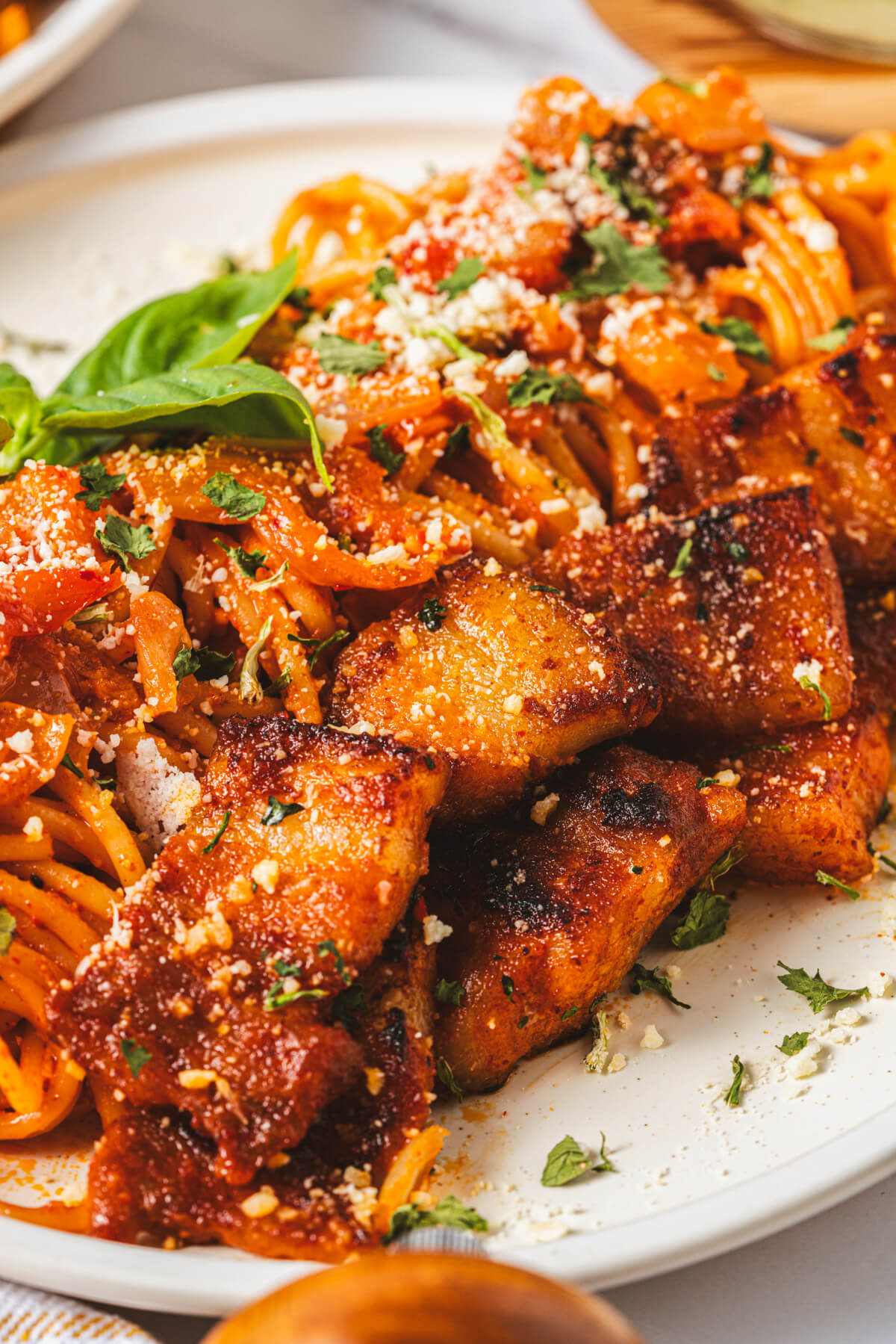 A white dinner plate with a serving of Kimchi Pasta with pork belly expertly rolled into a neat line then topped with Parmesan cheese and fresh basil leaves.
