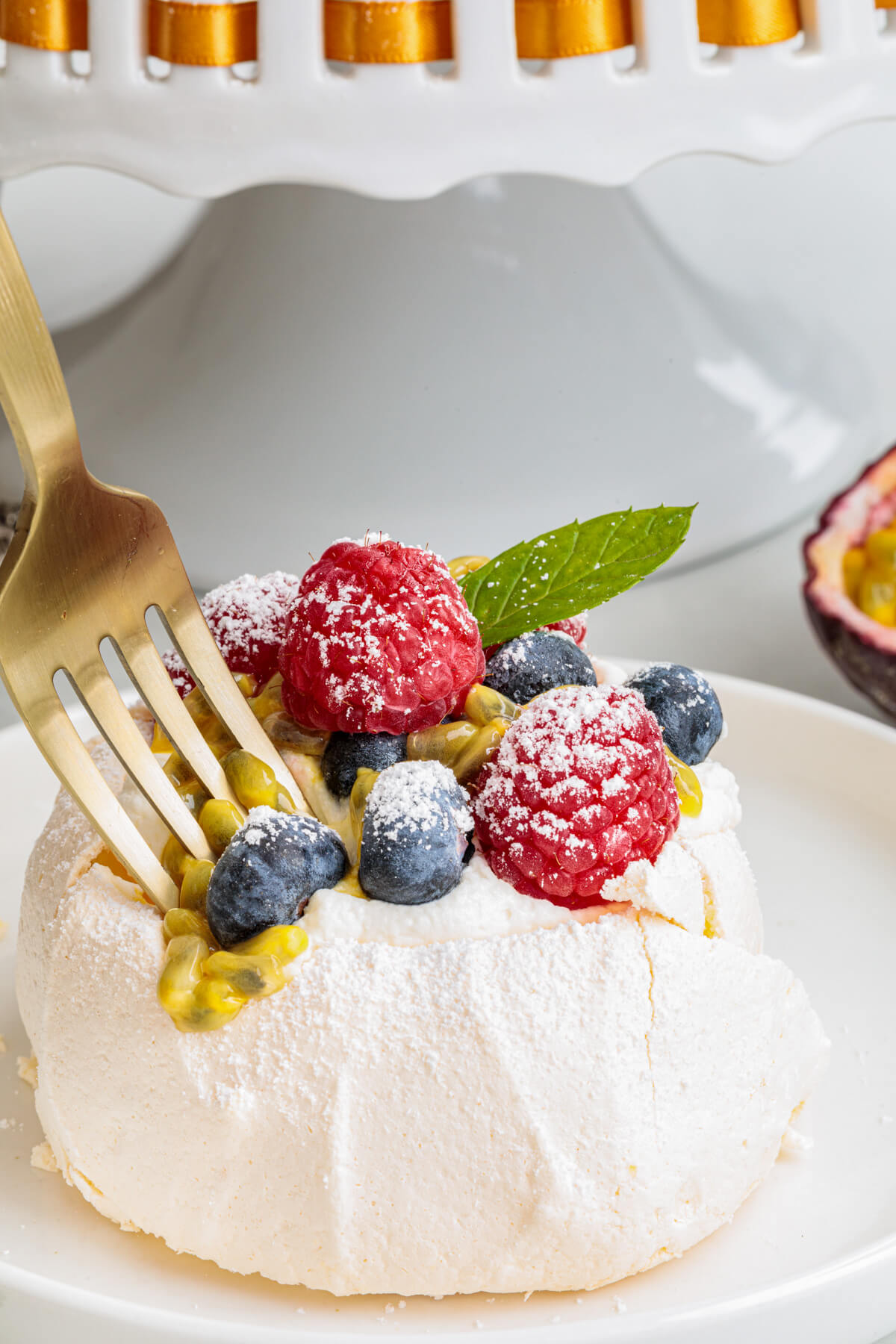 A fork digs into a Mini Pavlova topped with whipped cream, fresh berries, passionfruit pulp and fresh mint leaves on a serving platter.