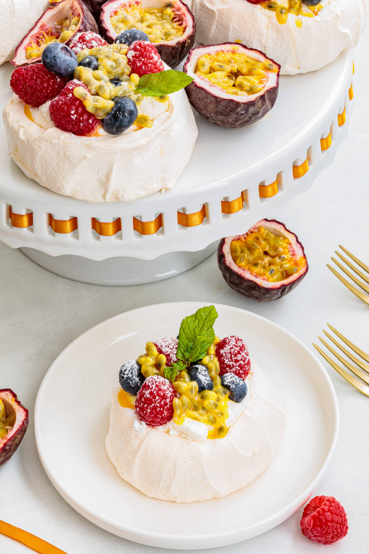One Mini Pavlova topped with whipped cream, fresh berries, passionfruit pulp and fresh mint leaves on a serving platter and another on a dessert plate.