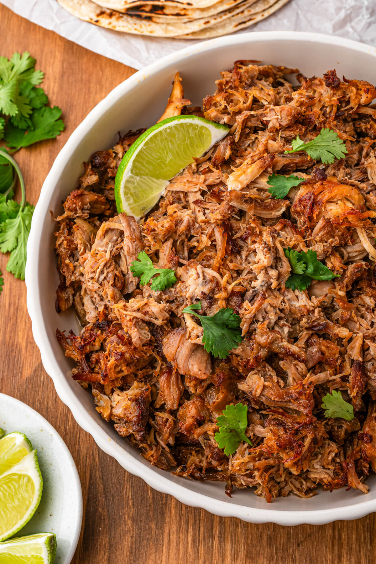 A white bowl of pork carnitas garnished with chopped cilantro and lime wedges.
