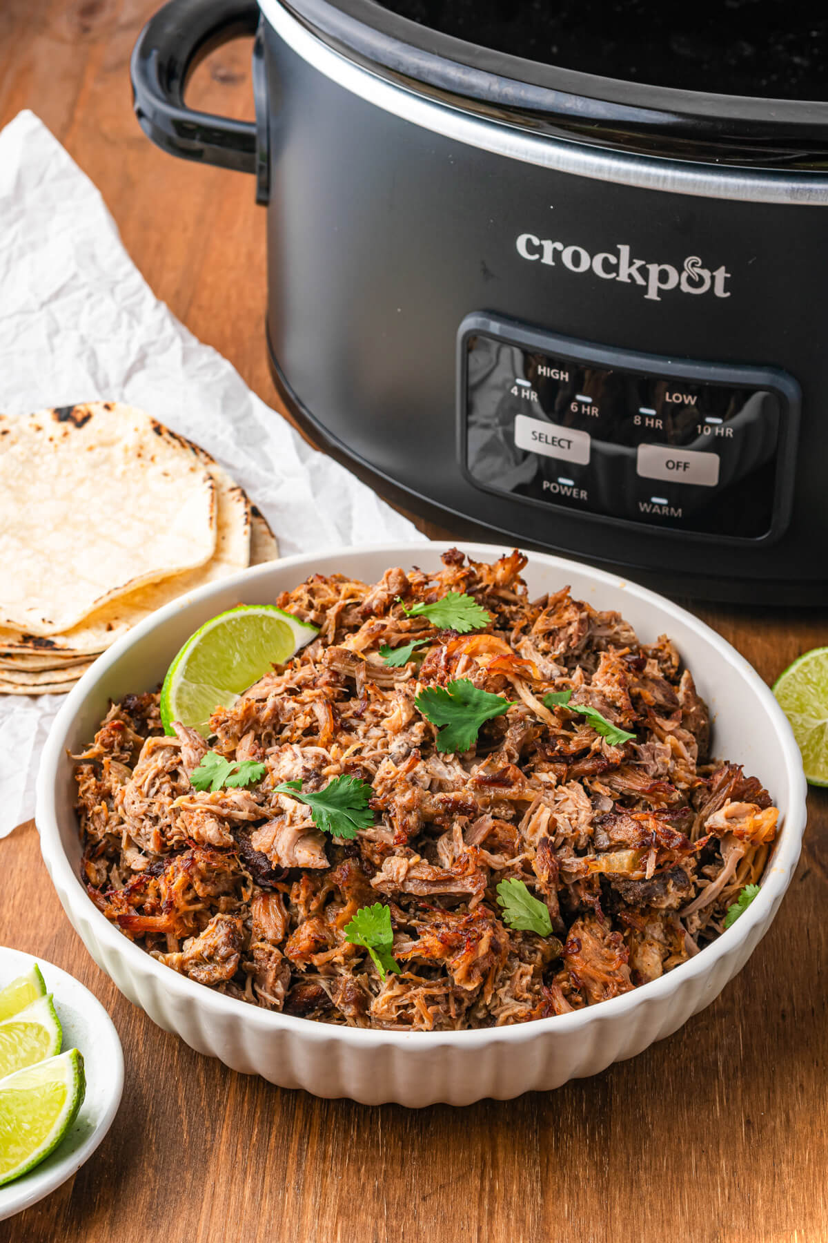 A white bowl of pork carnitas garnished with chopped cilantro and lime wedges next to a slow cooker.
