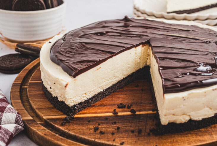 An almost whole Bailey's Cheesecake with a slice cut out sits on a wooden serving board surrounded by oreo cookies.