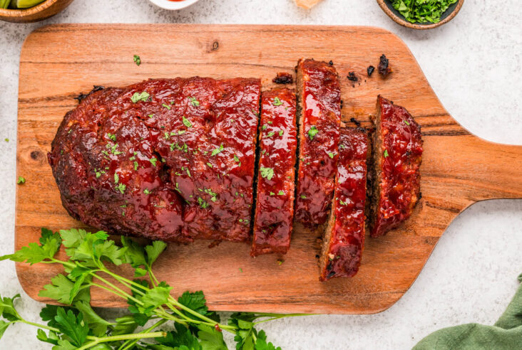 A half sliced shiny glazed smoked meatloaf on a wooden cutting board.