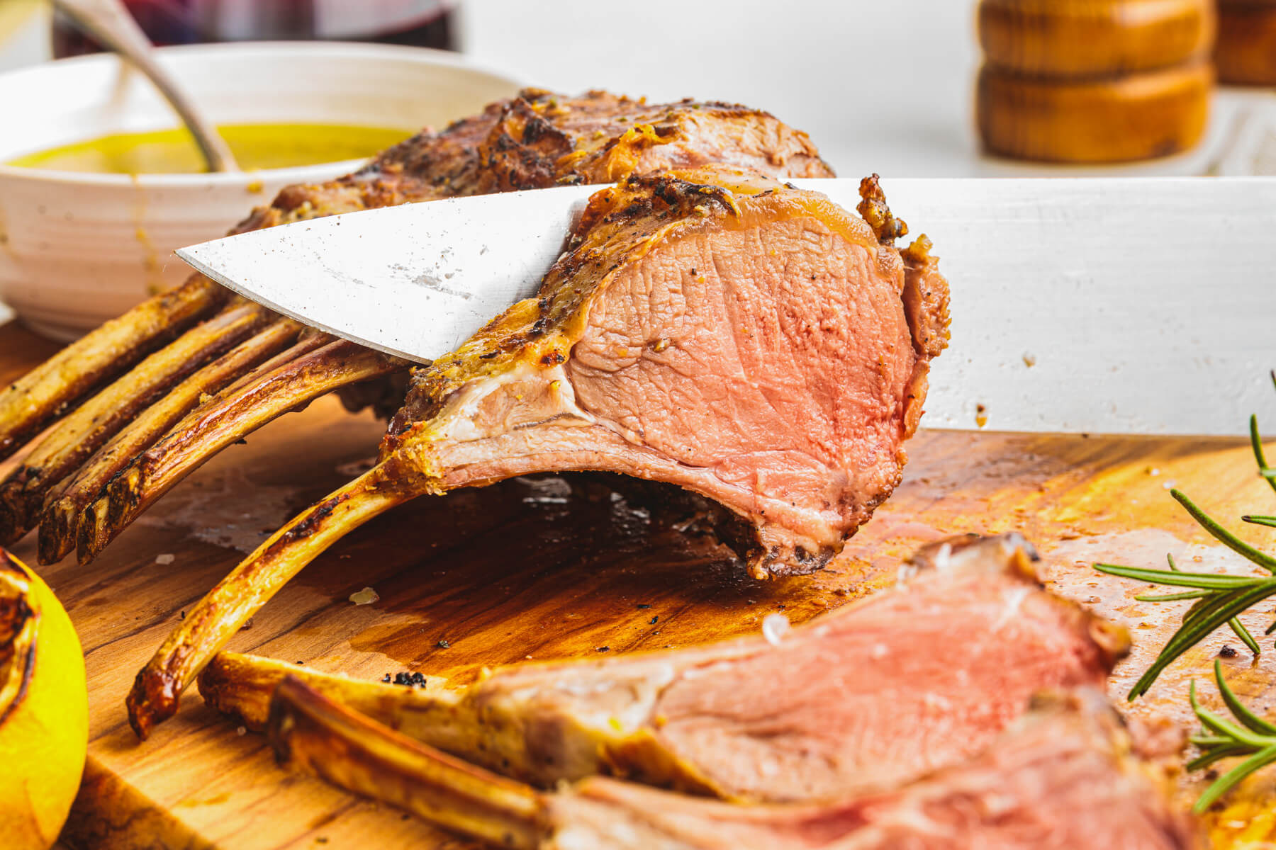One medium rare pink lamb chop being sliced from the whole rack on a wooden cutting board.