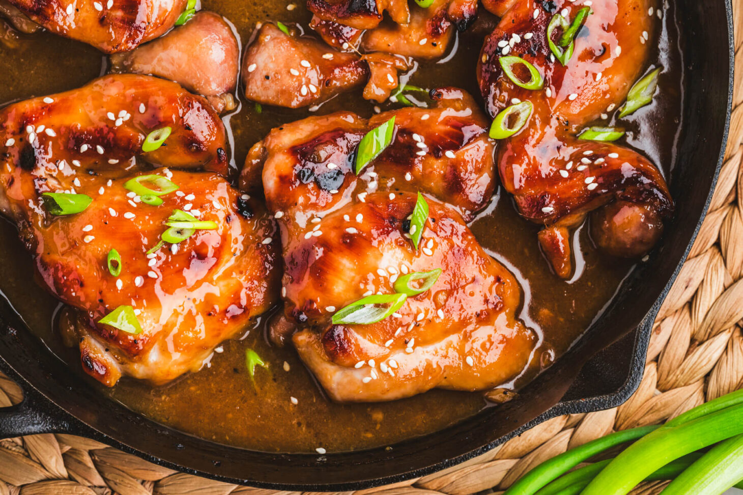 Glossy honey soy chicken thighs garnished with sliced green onions and white sesame seeds in a cast iron skillet.