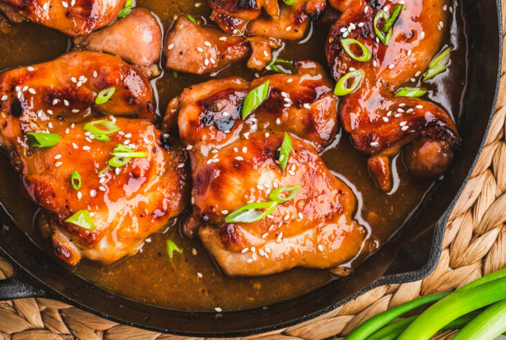 Glossy honey soy chicken thighs garnished with sliced green onions and white sesame seeds in a cast iron skillet.
