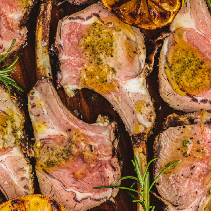 Several pink slices of lamb topped with rosemary dijon finishing oil on a wooden serving board.