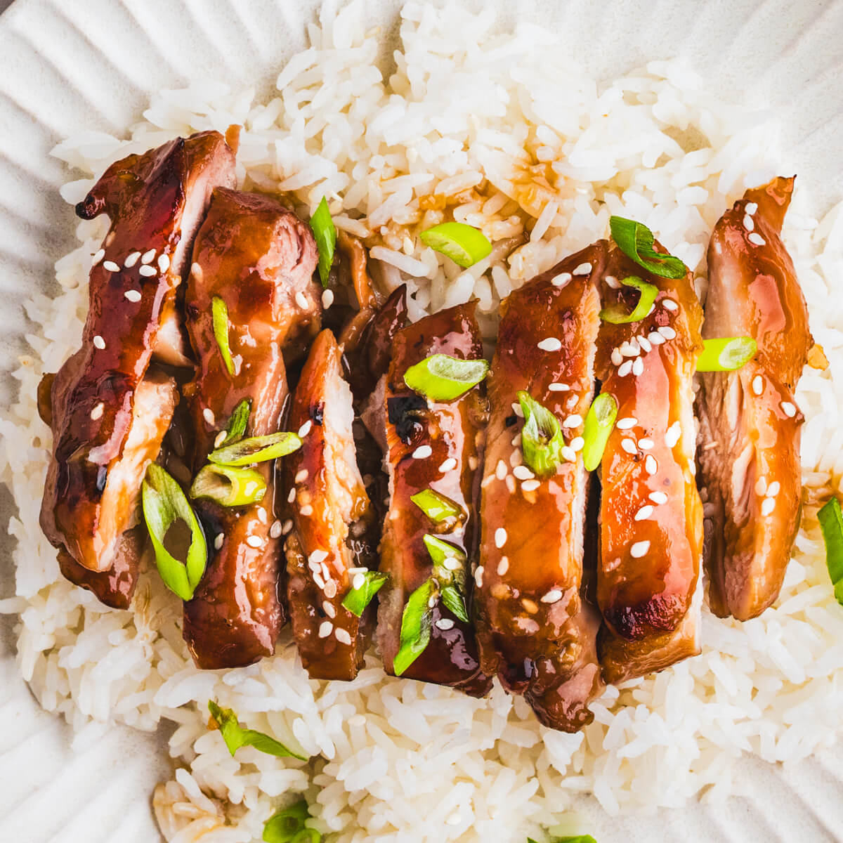 A sliced honey soy chicken thigh sits on top of a bed of rice garnished with sliced green onions and white sesame seeds.