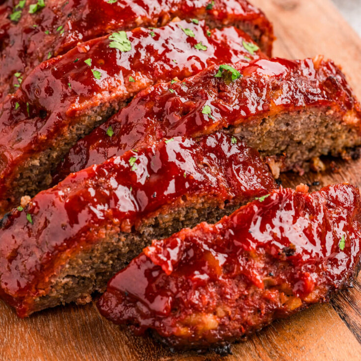 A half sliced shiny glazed smoked meatloaf on a wooden cutting board.
