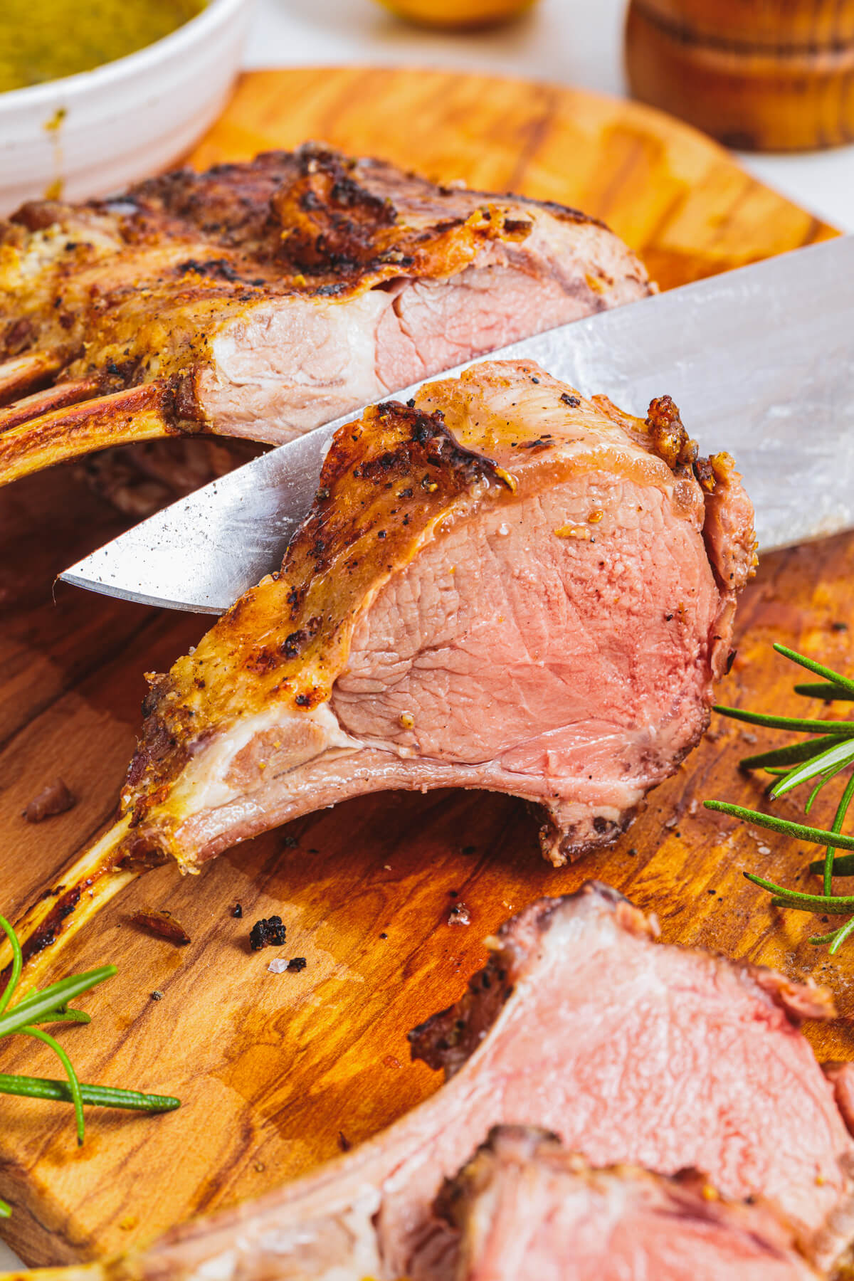 One medium rare pink lamb chop being sliced from the whole rack on a wooden cutting board.