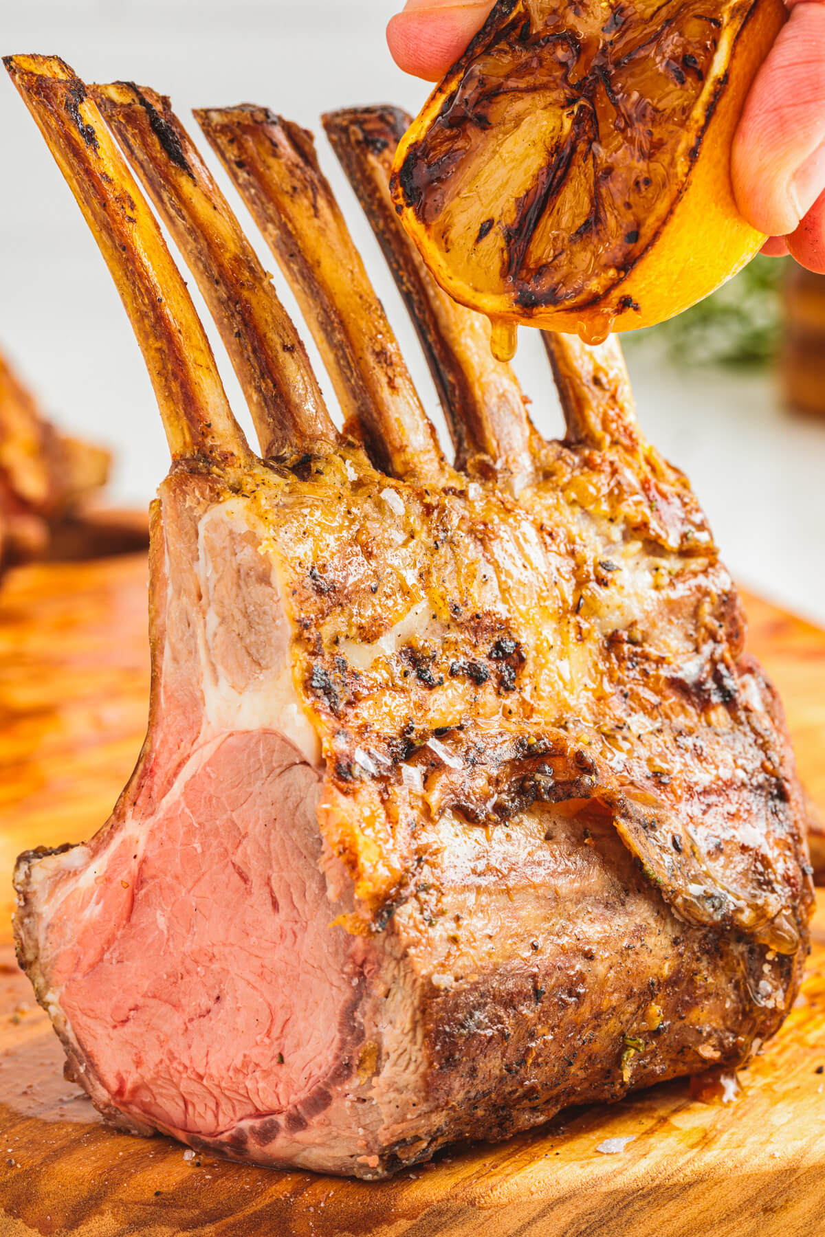 A perfectly grilled rack of lamb with the first slice missing being drizzled with lemon juice just before carving on a wooden cutting board.