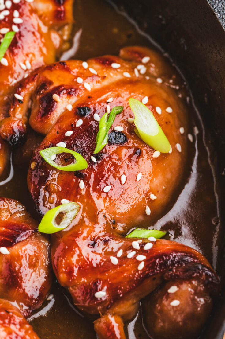 Close up of a single glossy honey soy chicken thigh garnished with sliced green onions and white sesame seeds.