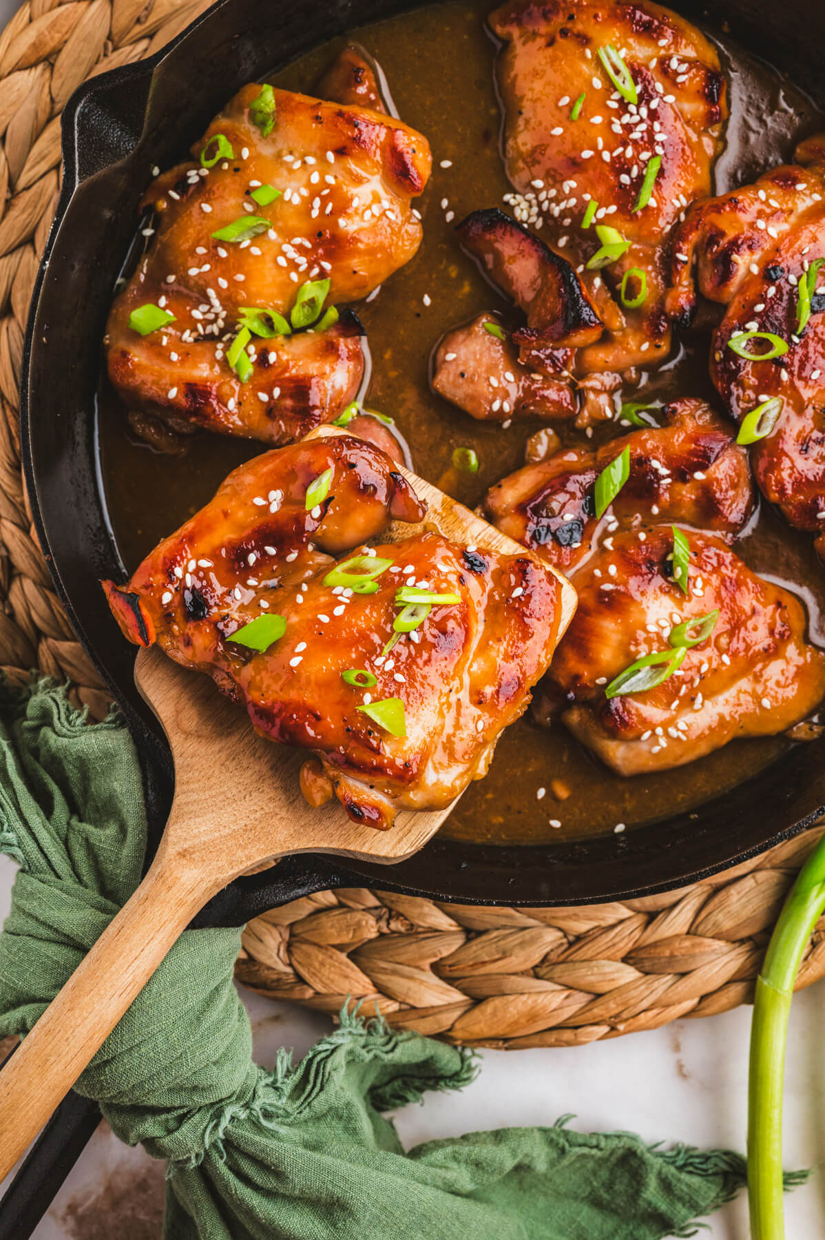 A wooden spoon serves a glossy honey soy chicken thigh from a black skillet full of chicken.