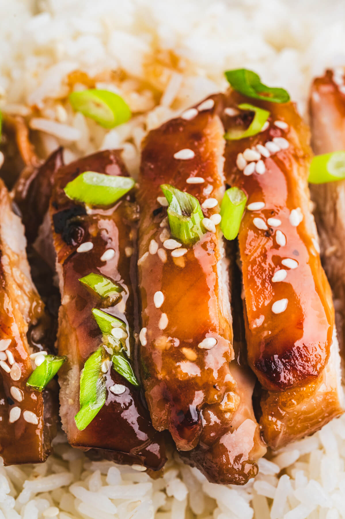 A sliced honey soy chicken thigh sits on top of a bed of rice garnished with sliced green onions and white sesame seeds.