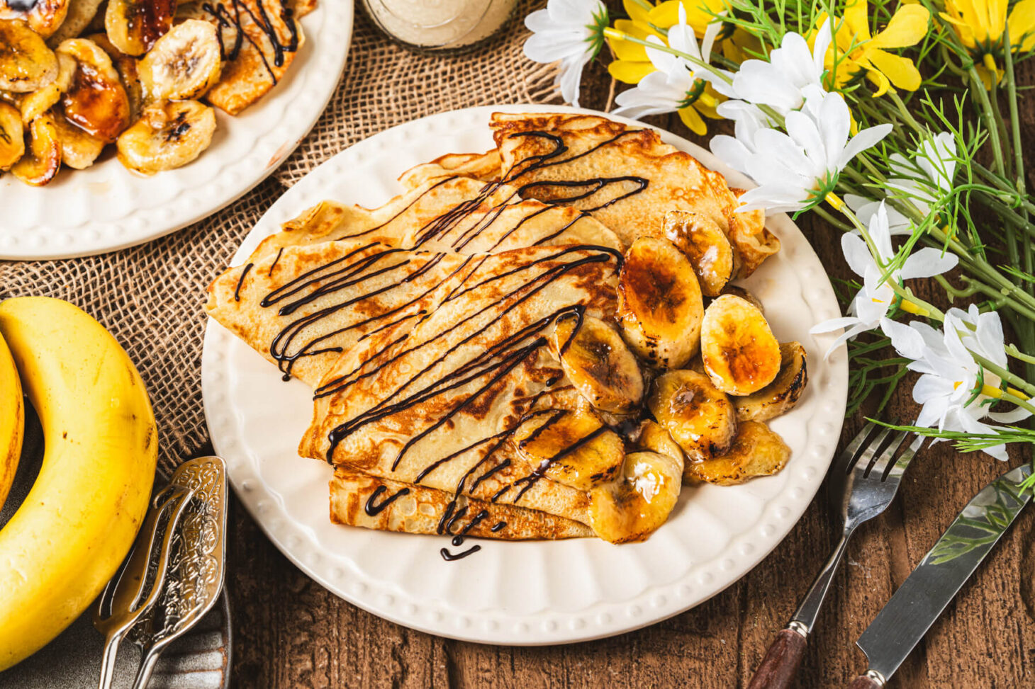 A white plate filled with folded Banana crepes drizzled in chocolate sauce and caramelized bananas.