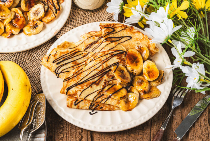 A white plate filled with folded Banana crepes drizzled in chocolate sauce and caramelized bananas.