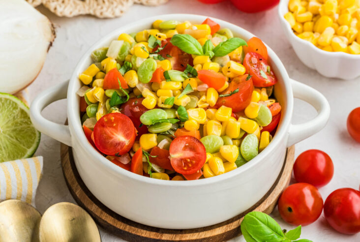A white serving dish filled with vibrant yellow corn succotash featuring green lima beans, red bell peppers, cherry tomatoes, and fresh basil.