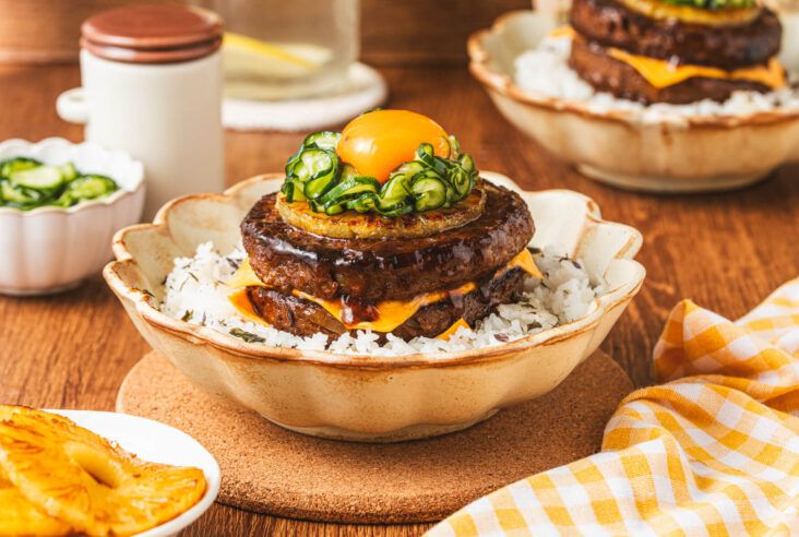 A two layer Japanese Hambagu steak dish piled high with American cheese, grilled pineapple, picked cucumber and topped with an egg yolk on a bed of seasoned rice.