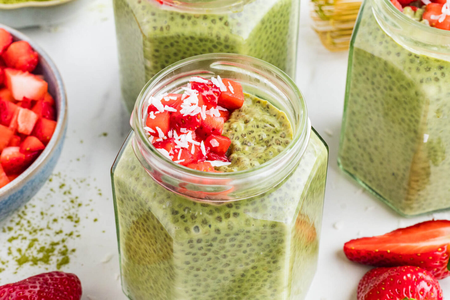Four jars of vibrant green Match Chia Pudding topped with sliced strawberries and coconut flakes.