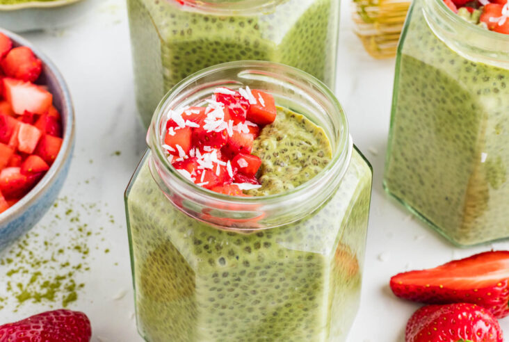 Four jars of vibrant green Match Chia Pudding topped with sliced strawberries and coconut flakes.