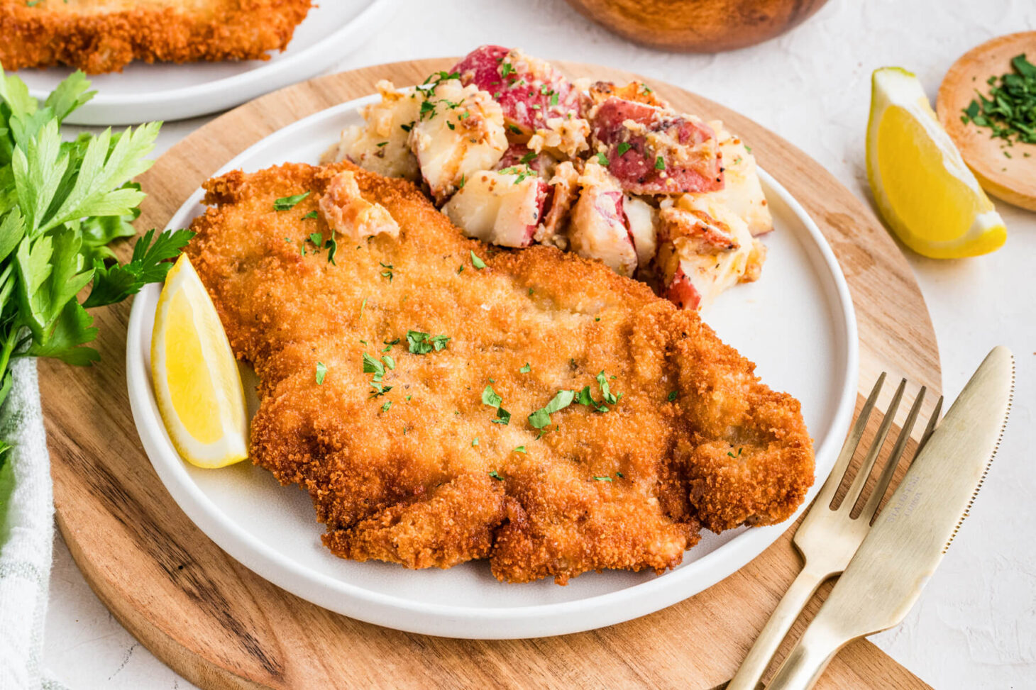 A golden brown fried pork schnitzel on a plate with a lemon wedge and a side of potato salad.