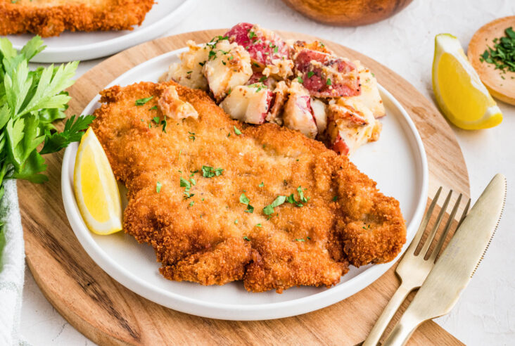 A golden brown fried pork schnitzel on a plate with a lemon wedge and a side of potato salad.
