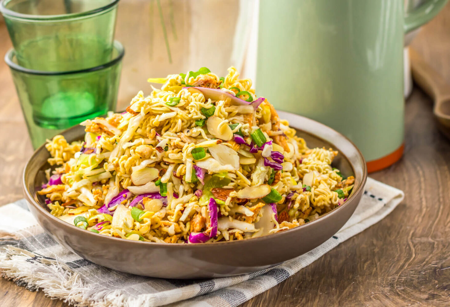 A brown bowl piled high with ramen noodle salad featuring pulled rotisserie chicken, coleslaw mix, sesame seeds, and green onions.