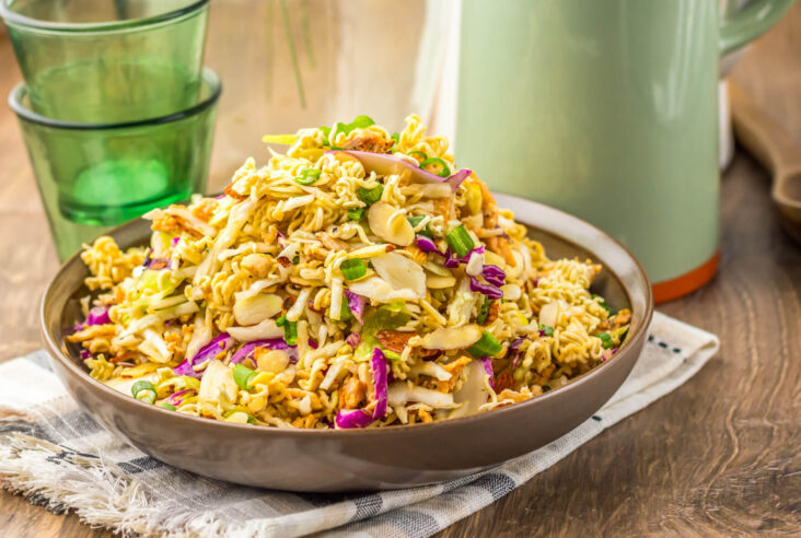 A brown bowl piled high with ramen noodle salad featuring pulled rotisserie chicken, coleslaw mix, sesame seeds, and green onions.