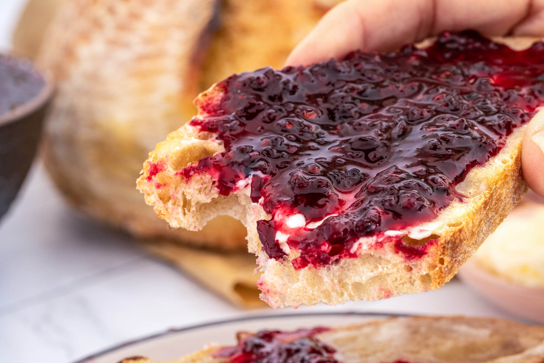 A hand holds a slice of sourdough toast topped with butter and blackberry jam.