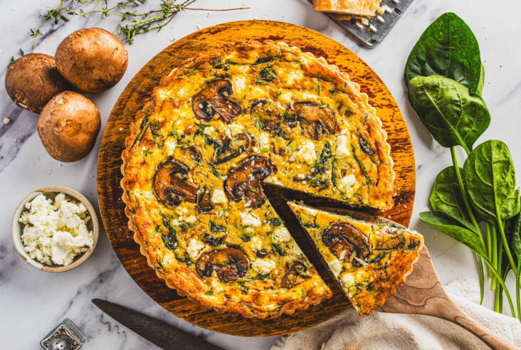 A slice of Spinach Mushroom Quiche in a golden baked crust on a wooden spatula resting above the whole quiche.