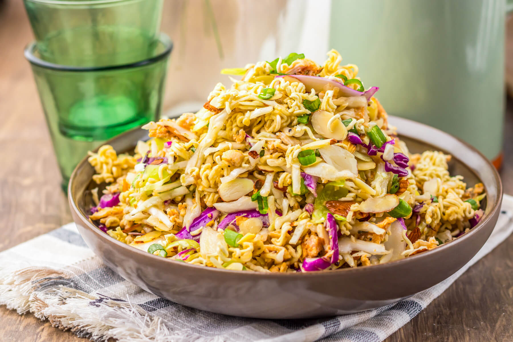 A brown bowl piled high with ramen noodle salad featuring pulled rotisserie chicken, coleslaw mix, sesame seeds, and green onions.