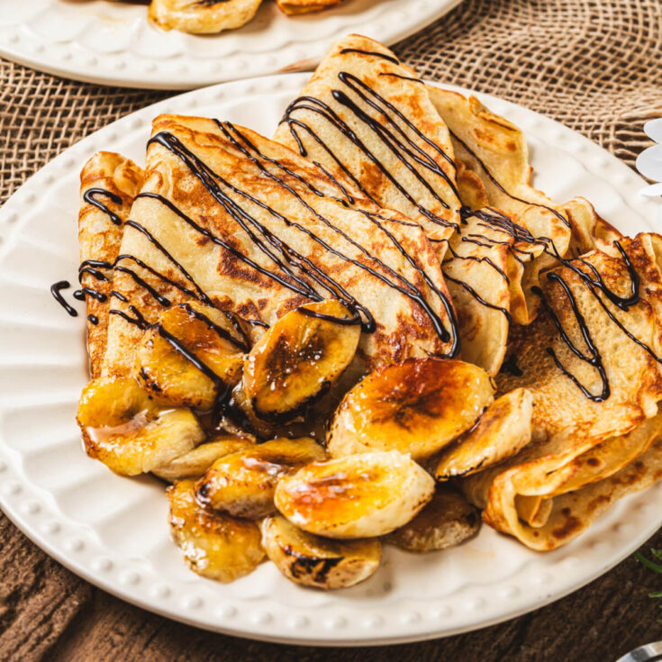 A white plate filled with folded Banana crepes drizzled in chocolate sauce and caramelized bananas.