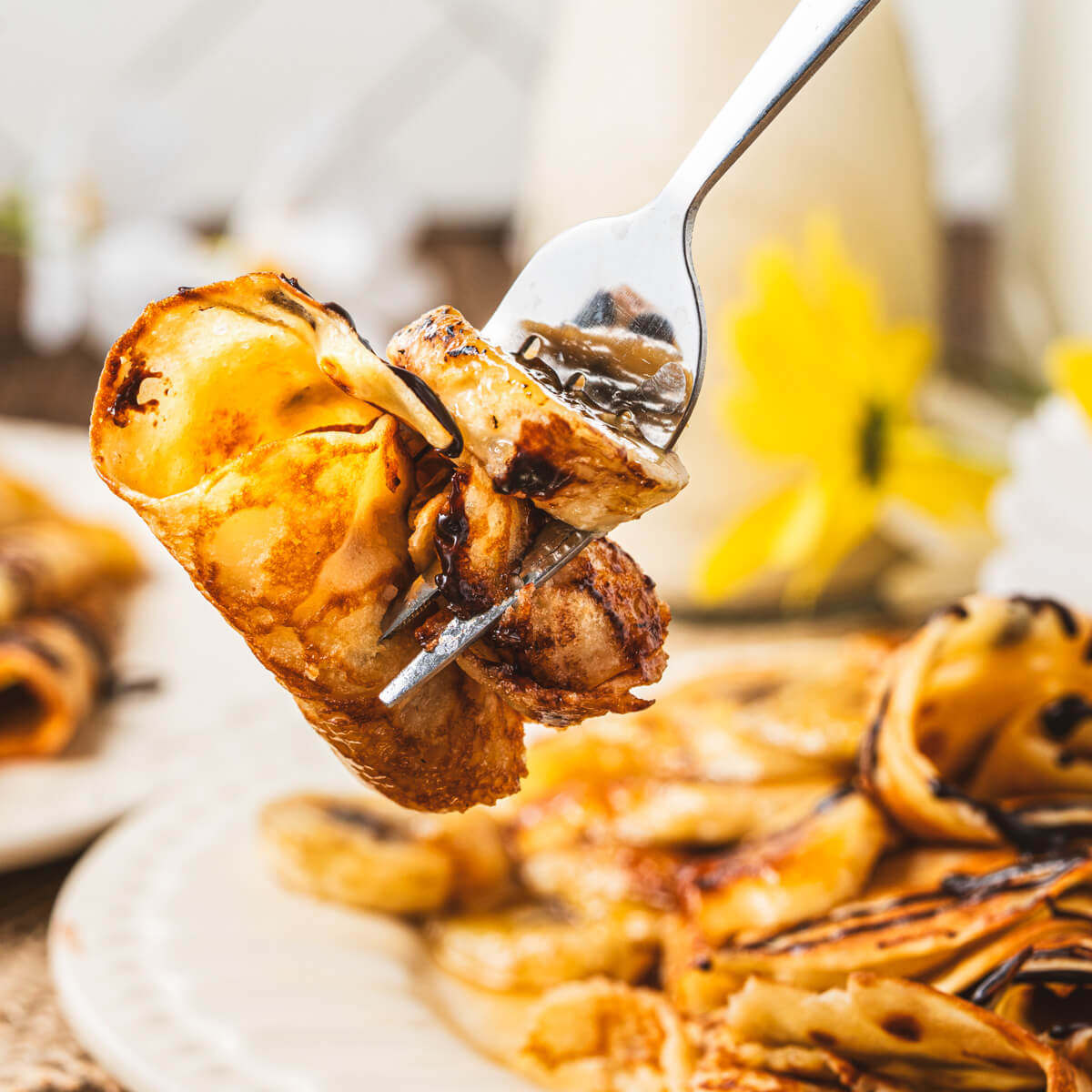 A fork holding a folded banana crepe topped with a caramelilzed banana and chocolate sauce.