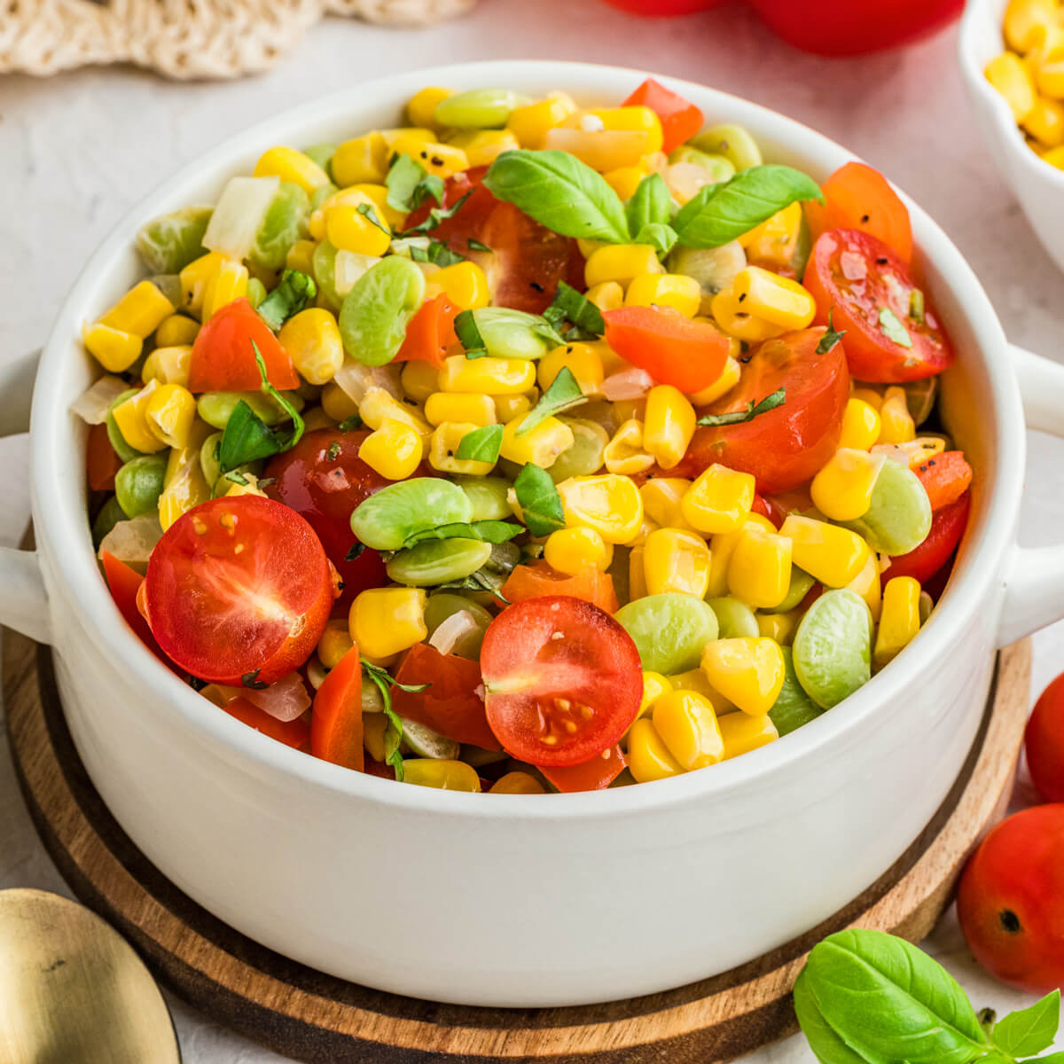 A white serving dish filled with vibrant yellow corn succotash featuring green lima beans, red bell peppers, cherry tomatoes, and fresh basil.