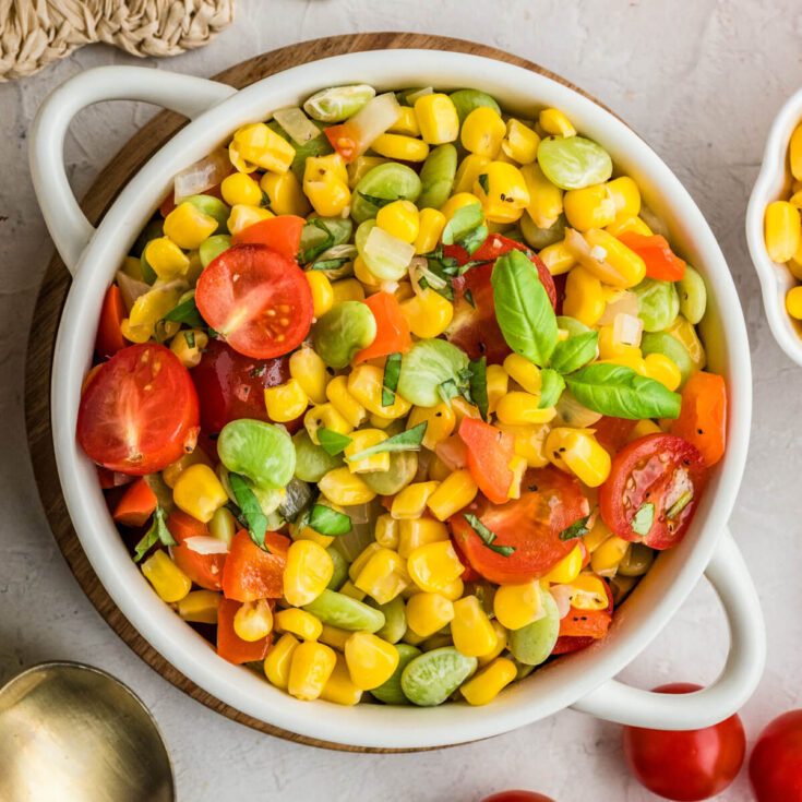 A white serving dish filled with vibrant yellow corn succotash featuring green lima beans, red bell peppers, cherry tomatoes, and fresh basil.
