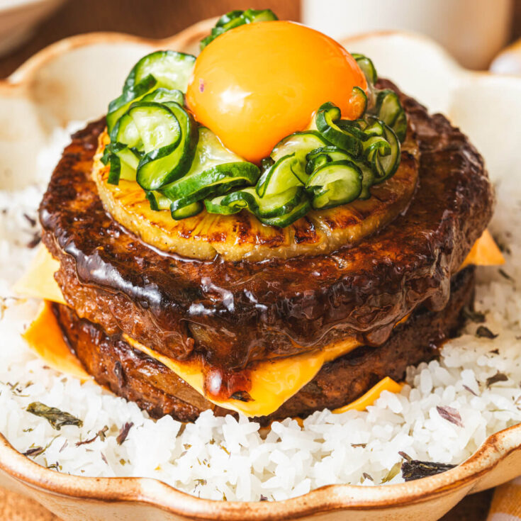 A two layer Japanese Hambagu steak dish piled high with American cheese, grilled pineapple, picked cucumber and topped with an egg yolk on a bed of seasoned rice.