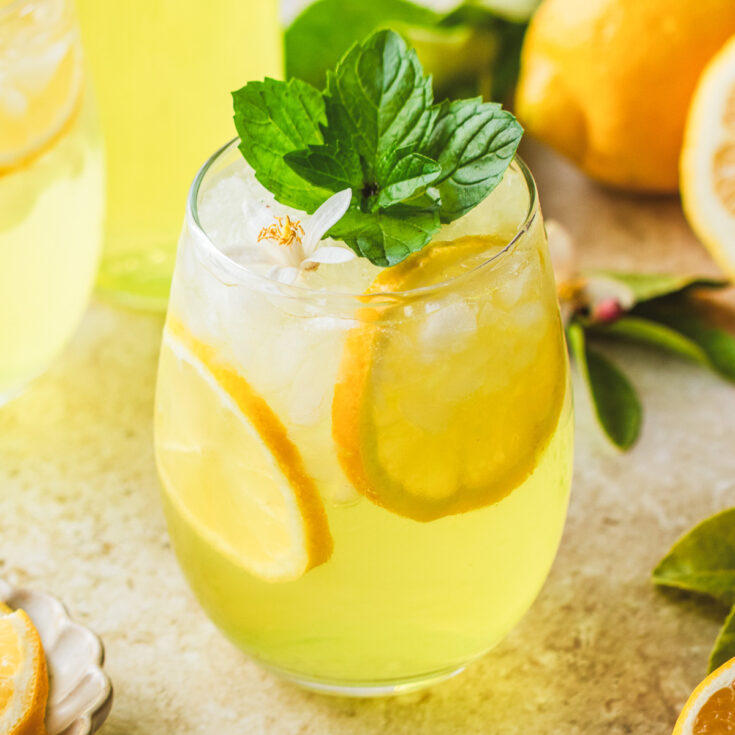 A sunny yellow cockail in a stemless wine glass garnished with fresh mint and lemon slices.