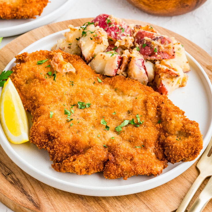 A golden brown fried pork schnitzel on a plate with a lemon wedge and a side of potato salad.