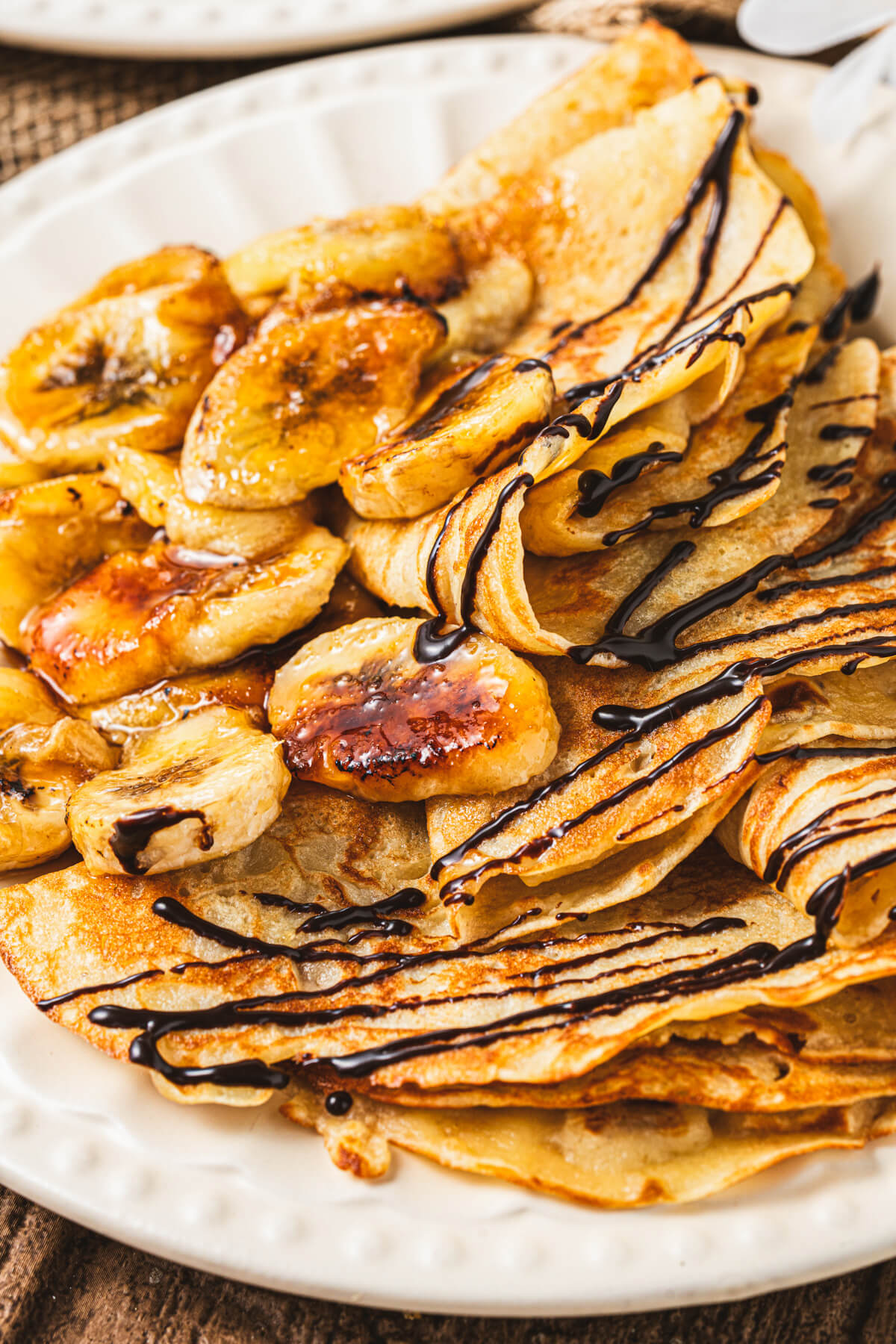A white plate filled with folded Banana crepes drizzled in chocolate sauce and caramelized bananas.