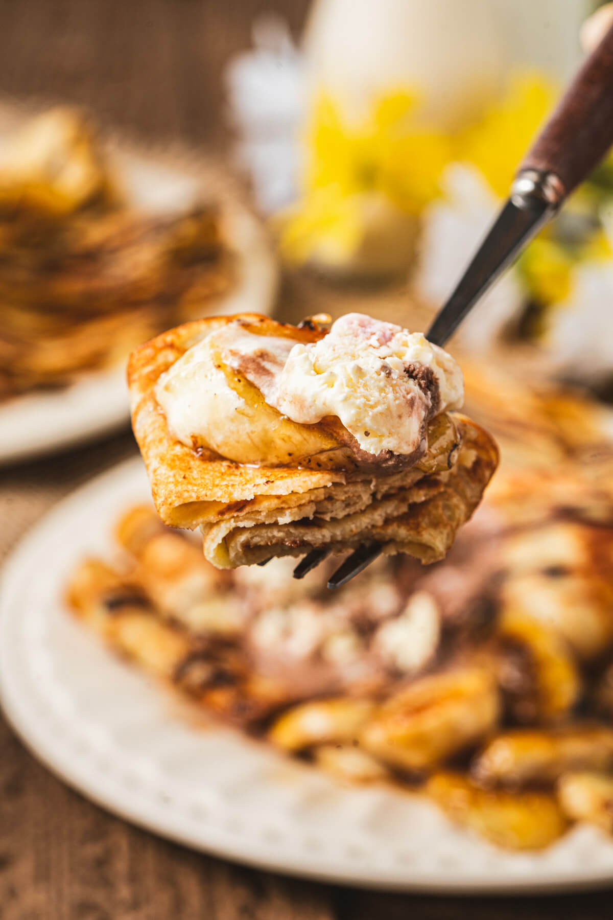 A fork holding a folded banana crepe topped with a bit of ice cream and chocolate sauce.