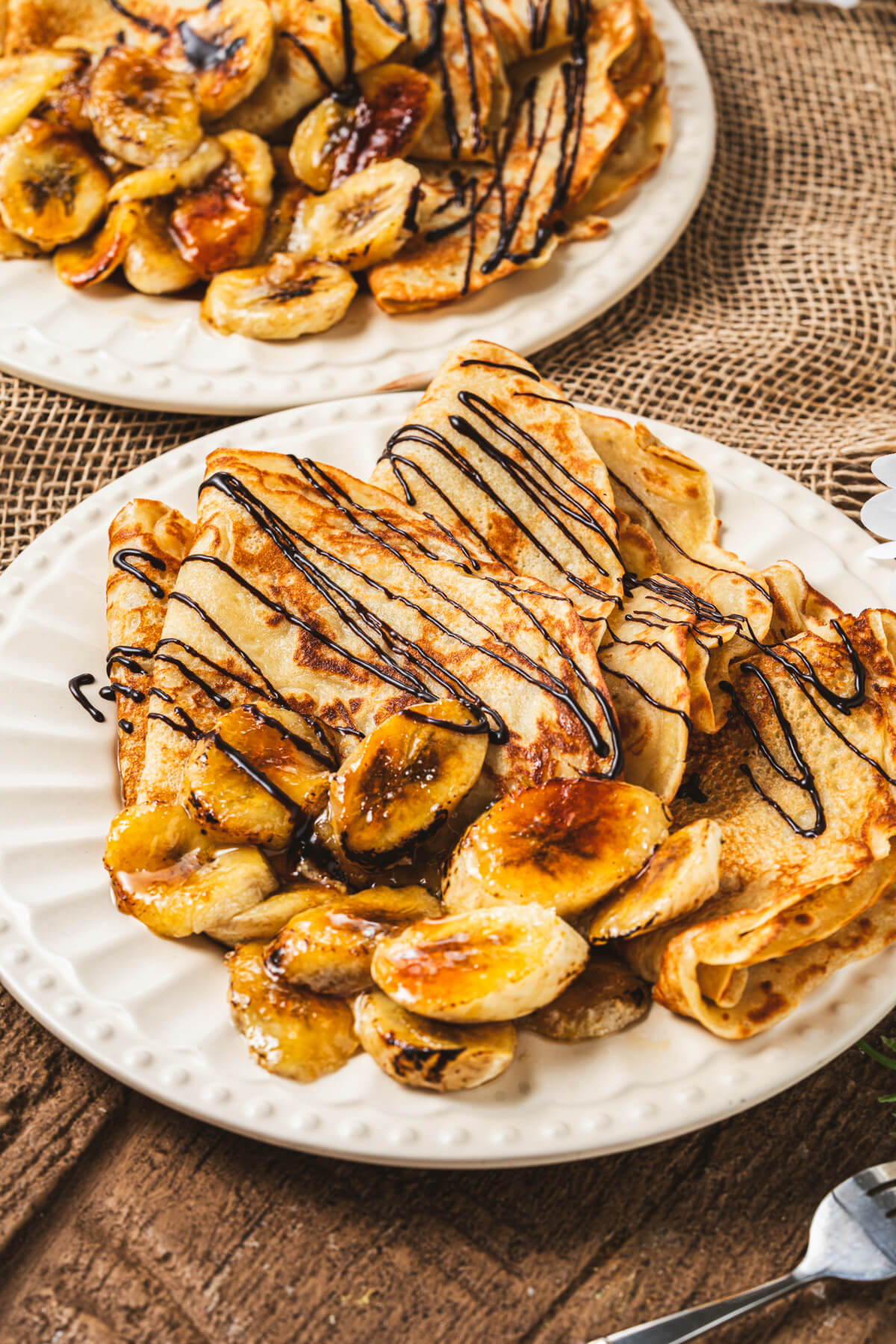 A white plate filled with folded Banana crepes drizzled in chocolate sauce and caramelized bananas.