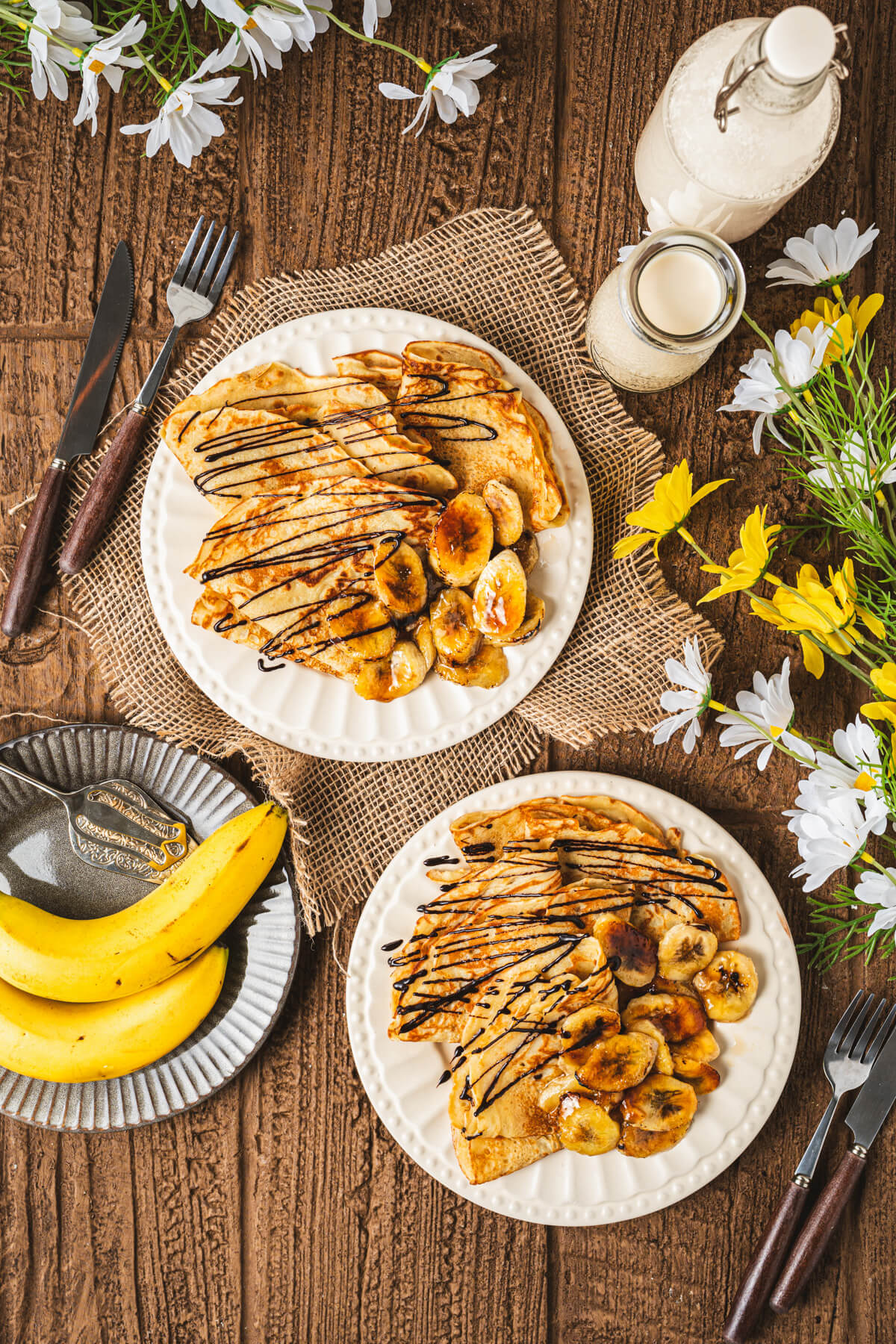 Two white plates filled with folded Banana crepes drizzled in chocolate sauce and caramelized bananas on a breakfast table.