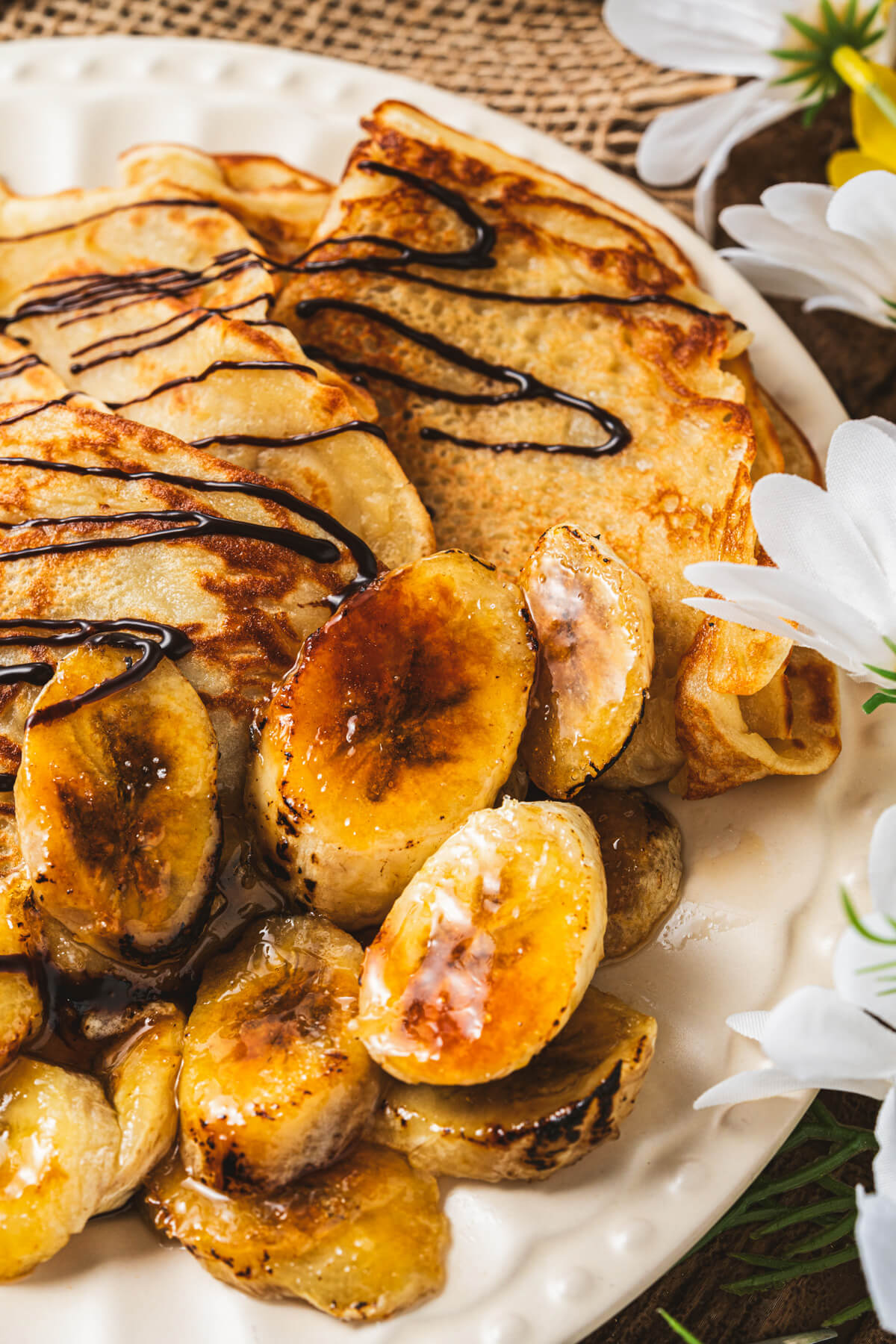 A white plate filled with folded Banana crepes drizzled in chocolate sauce and caramelized bananas.