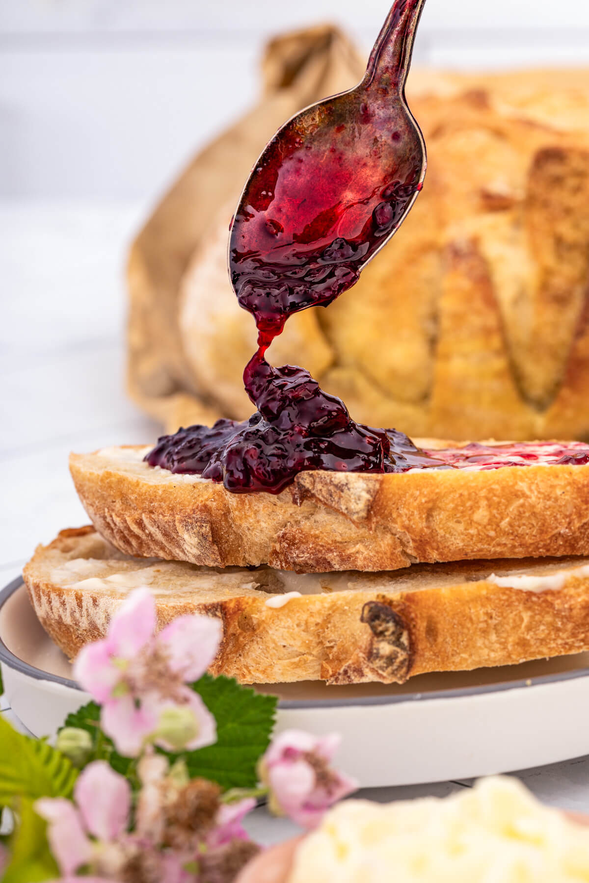 A spoon drizzles dark red blackberry jam on buttered sourdough toast.