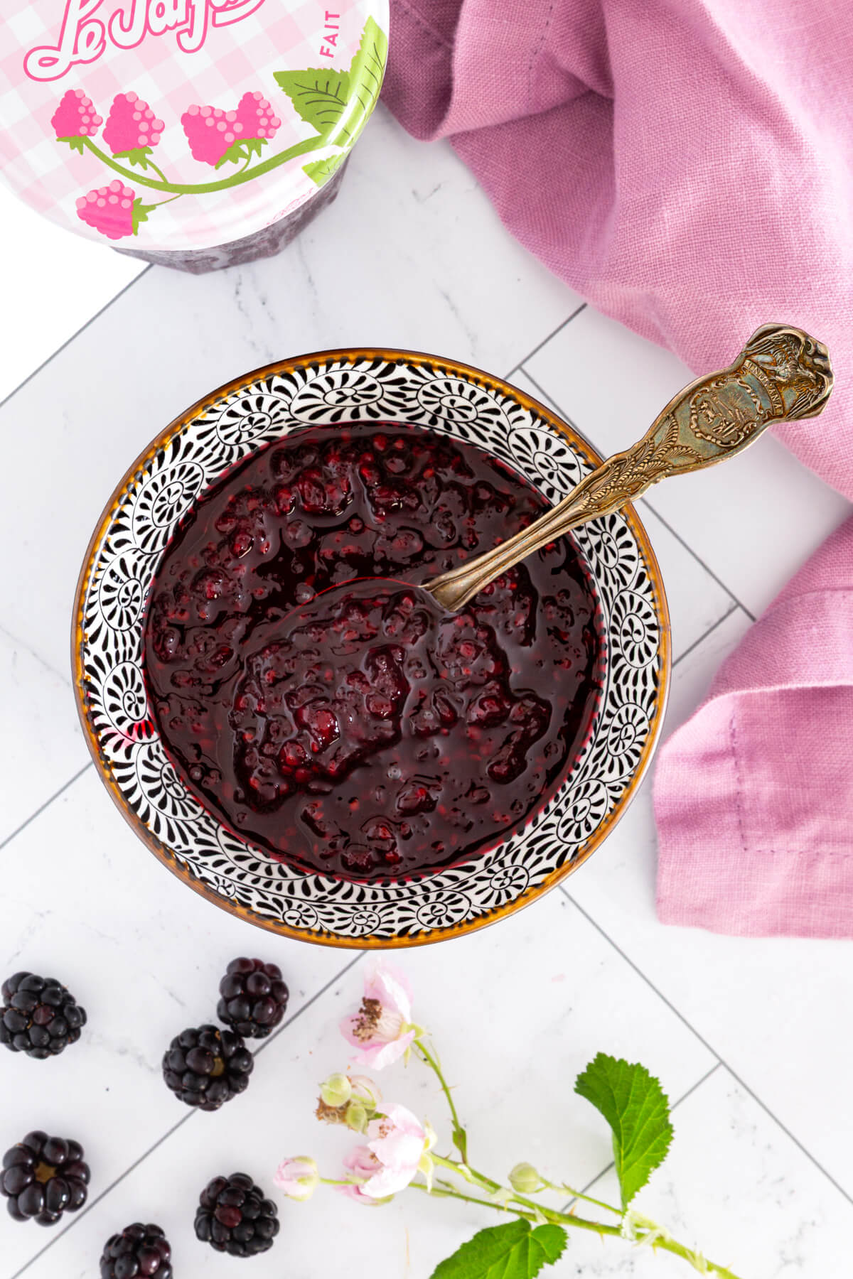 A spoon in a bowl of blackberry jam surrounded by blackberries and blackberry flowers.