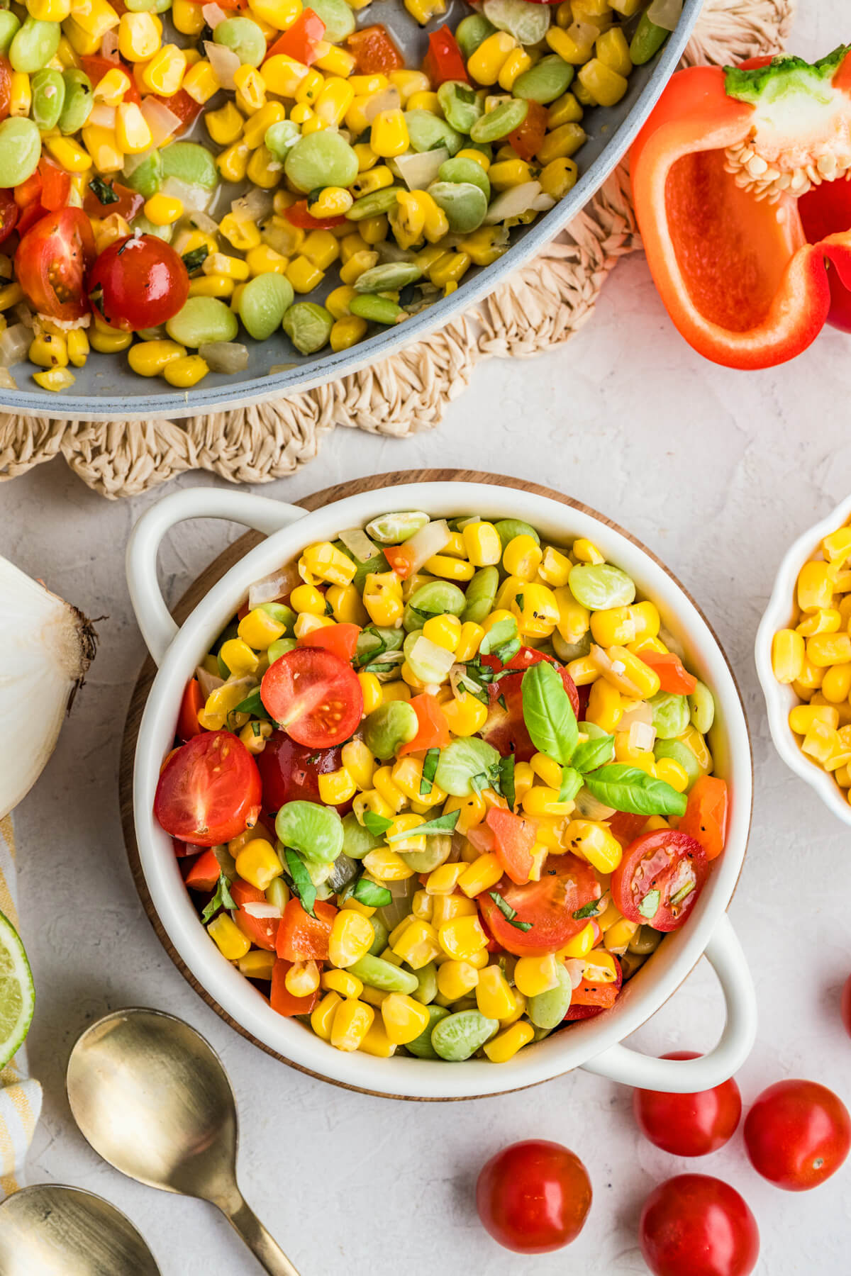 A white serving dish filled with vibrant yellow corn succotash featuring green lima beans, red bell peppers, cherry tomatoes, and fresh basil.