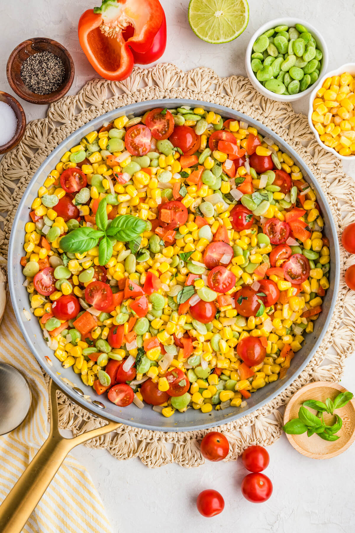 A skillet filled with vibrant yellow corn succotash featuring green lima beans, red bell peppers, cherry tomatoes, and fresh basil on a dinner table.